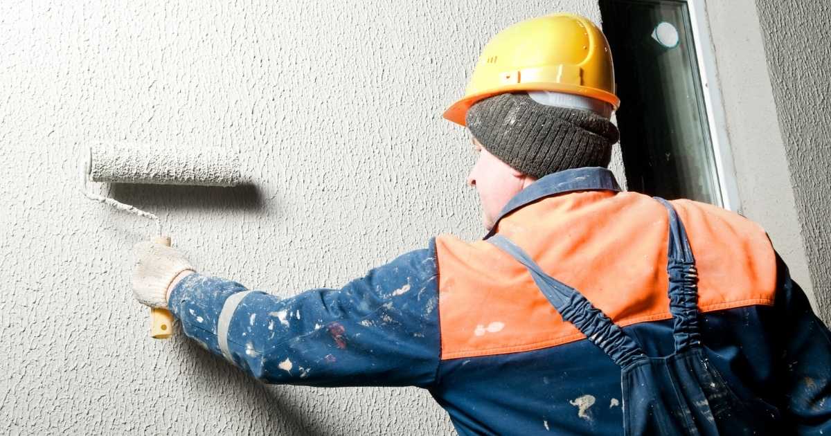Ouvrier en tenue de chantier en train de peindre une façade en crépi au rouleau.