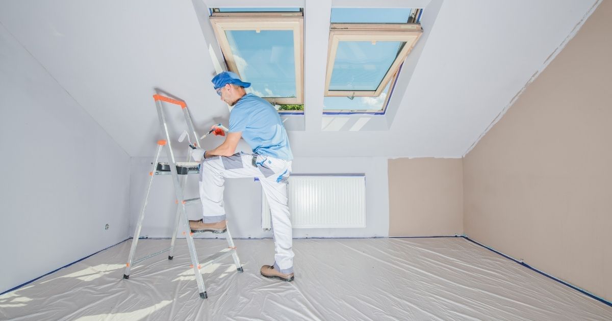 Peintre en bâtiment en train de peintre le plafond d'une pièce sous toiture avec une échelle et un pinceau.